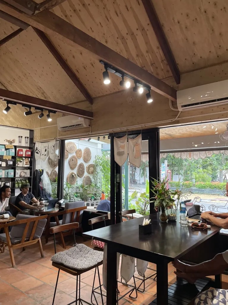 A bright wooden café interior with large windows, plants, and shared tables.