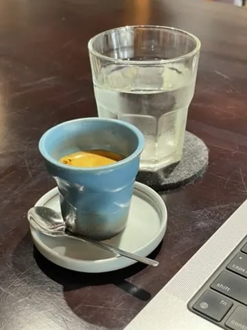 A blue ceramic espresso cup beside a laptop and a glass of water.