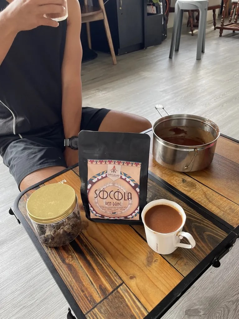 A hot cacao drink served beside cacao pieces and a bag of Vietnamese chocolate.