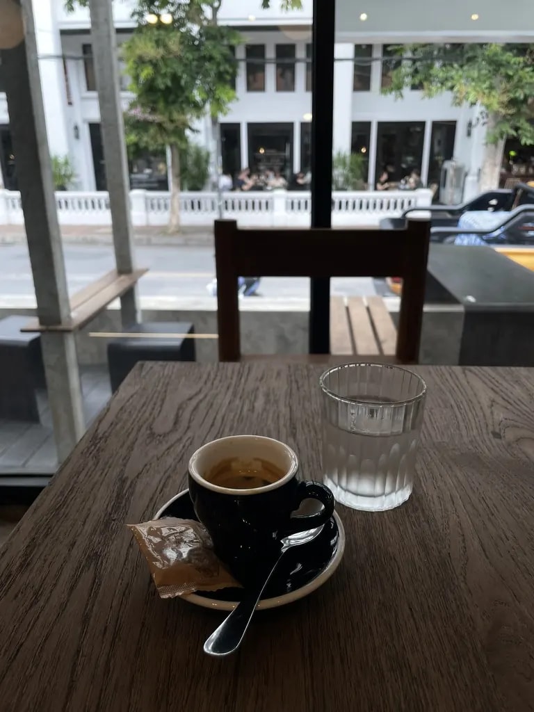 A black espresso cup and water on a wooden table facing a café window.