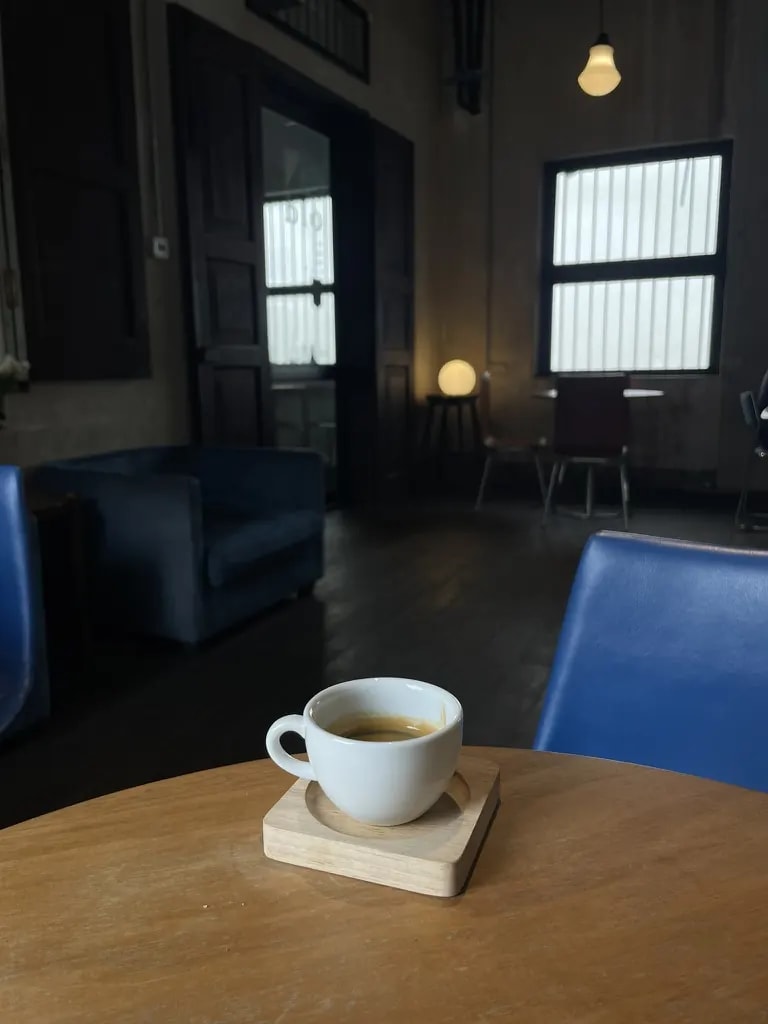 A white espresso cup in a dark café room with blue chairs.