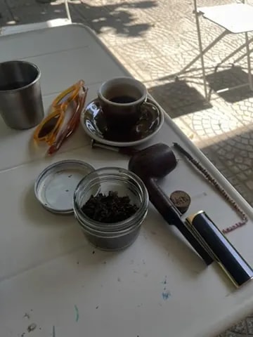 An espresso cup beside sunglasses, a pipe, and a small jar on an outdoor table.