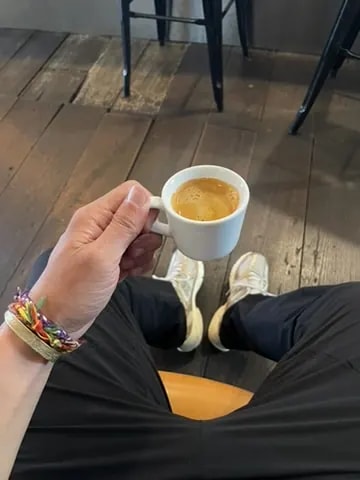 A hand holding a small white espresso cup above a wooden floor.
