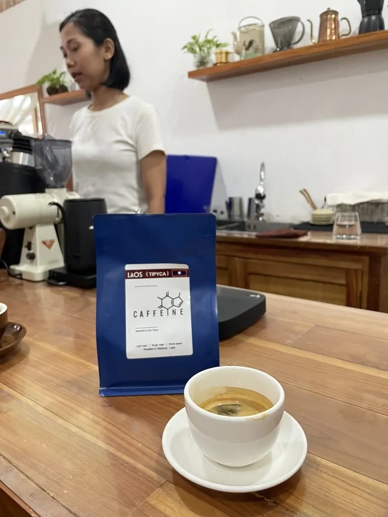 An espresso beside a bag of Lao coffee with a barista behind the counter.